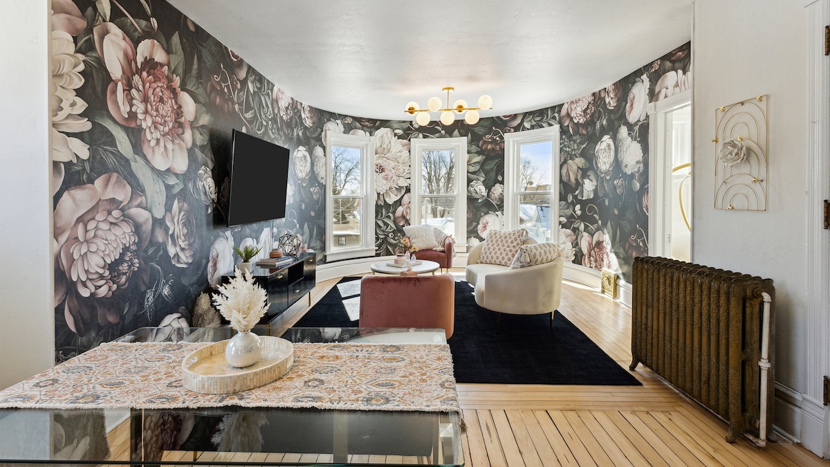 The living area features a floral wallpaper design, enhancing the space's character. Natural light fills the room through large curved windows, illuminating a stylish seating arrangement with a sofa and chairs. A decorative table is positioned in front, while a vintage radiator adds charm.