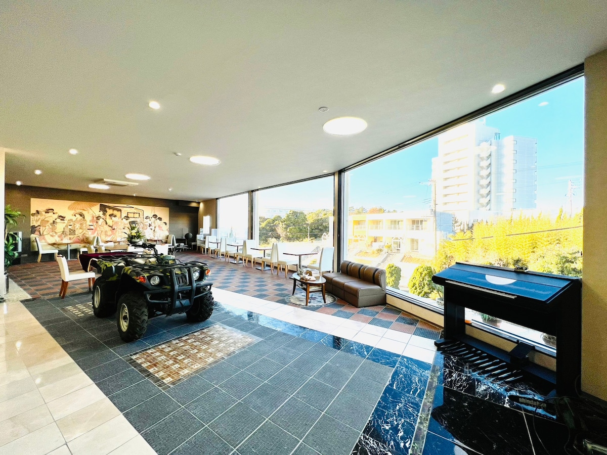 The lobby area features large windows allowing natural light to fill the space. A seating arrangement, including a brown sofa, is positioned near an elegant black piano. An ATV is displayed prominently on the tiled floor, showcasing a mix of textures and comfort.