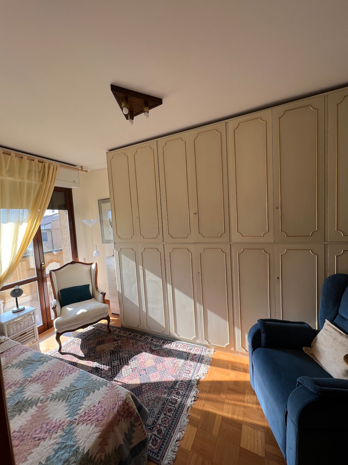 A bright room features a spacious bed draped with a colorful quilt, complemented by a blue armchair. Tall closets with elegant detailing maximize storage. Natural light pours in through a large window covered with sheer curtains, enhancing the inviting atmosphere.