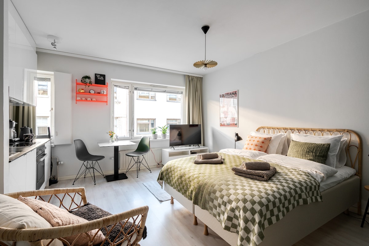 A combined living and sleeping area is highlighted with a spacious bed adorned with a checkered blanket. A small dining table with chairs is located beside a window, alongside a TV and a decorative shelf. A comfortable chair adds to the inviting layout.