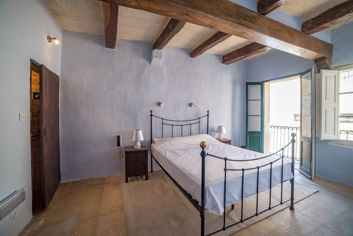 A spacious bedroom features a wrought iron bed with an unmade white mattress, complemented by two bedside lamps. Exposed wooden beams line the ceiling, and a balcony door opens to a balcony with slatted shutters, allowing natural light to fill the room.