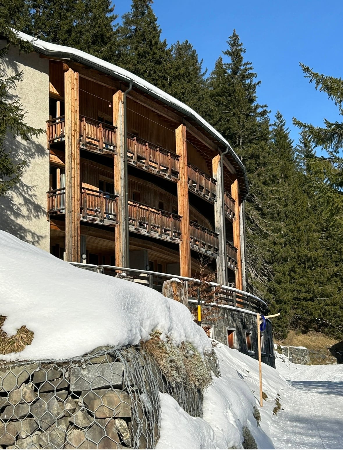 The exterior of a multi-level building is showcased, featuring wooden balconies that overlook a snowy landscape. Pine trees surround the property, and a gentle slope leads to the entrance. Snow blankets the ground, contributing to the serene alpine setting.