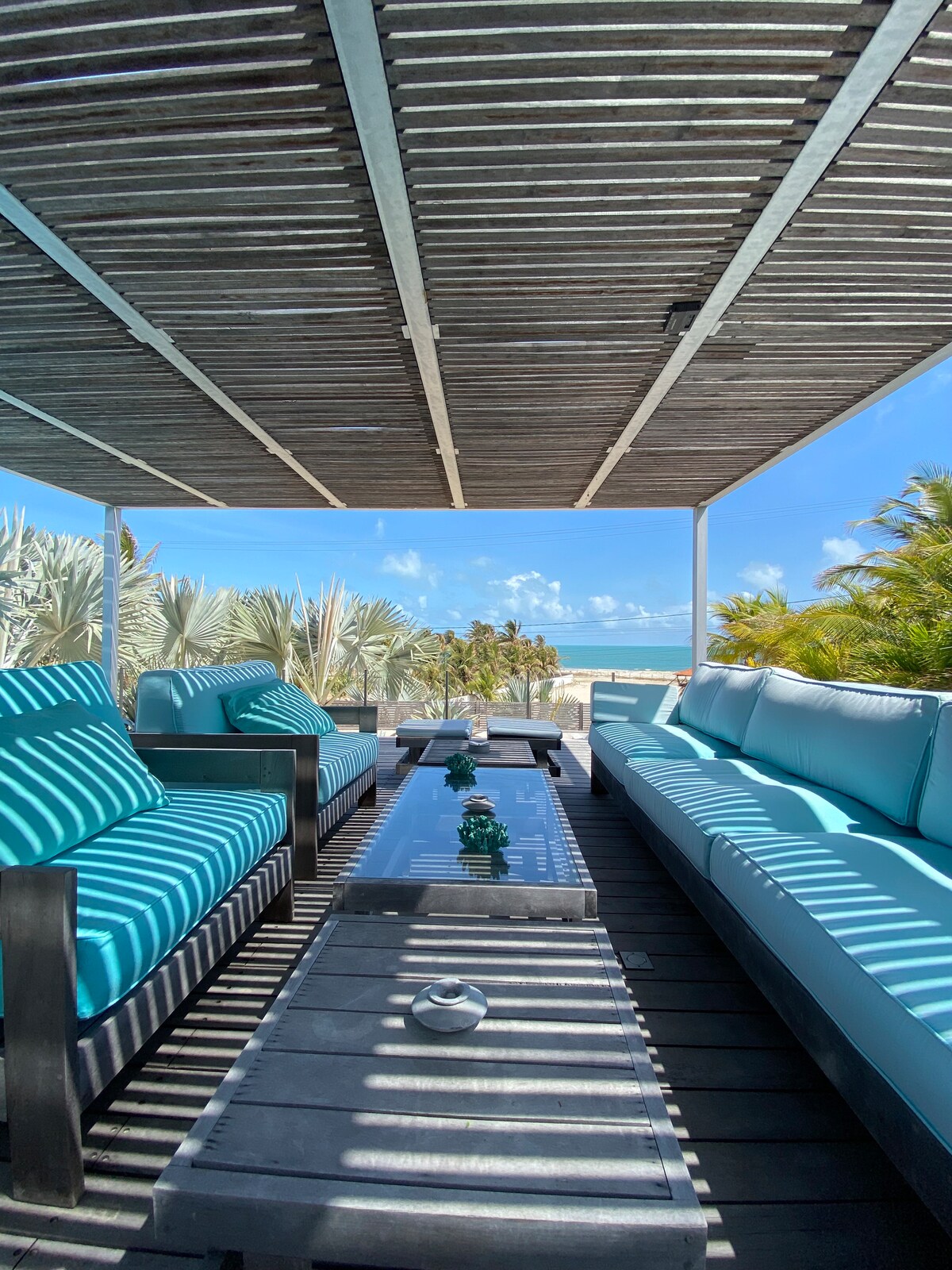 A shaded seating area features light blue cushioned sofas and a long wooden table, highlighting a calm outdoor space. Lush greenery and palm trees can be seen in the background, framing a clear view of the beach and sky.