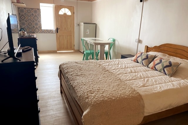 A spacious room is featured, equipped with a comfortable bed adorned with a textured blanket. A modern dining table with green chairs is positioned next to a wooden dresser and a television, while a door opens to the outside, providing natural light.