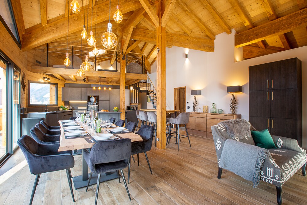 An open dining area features a large rectangular table set for 14 guests, with dark upholstered chairs positioned around it. A cozy lounge area is visible, with a comfortable chair and stylish lighting fixtures hanging from the vaulted wooden ceiling, enhancing the spacious design.