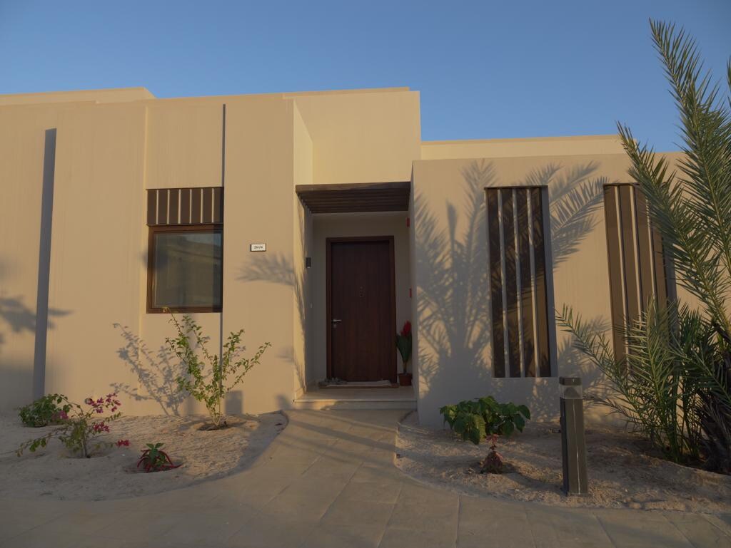 The entrance of a villa is framed by sandy pathways and landscaped greenery. A large wooden door is set into a beige façade, complemented by vertical wooden slats and a large window. Shadows from nearby plants add texture to the entrance area.
