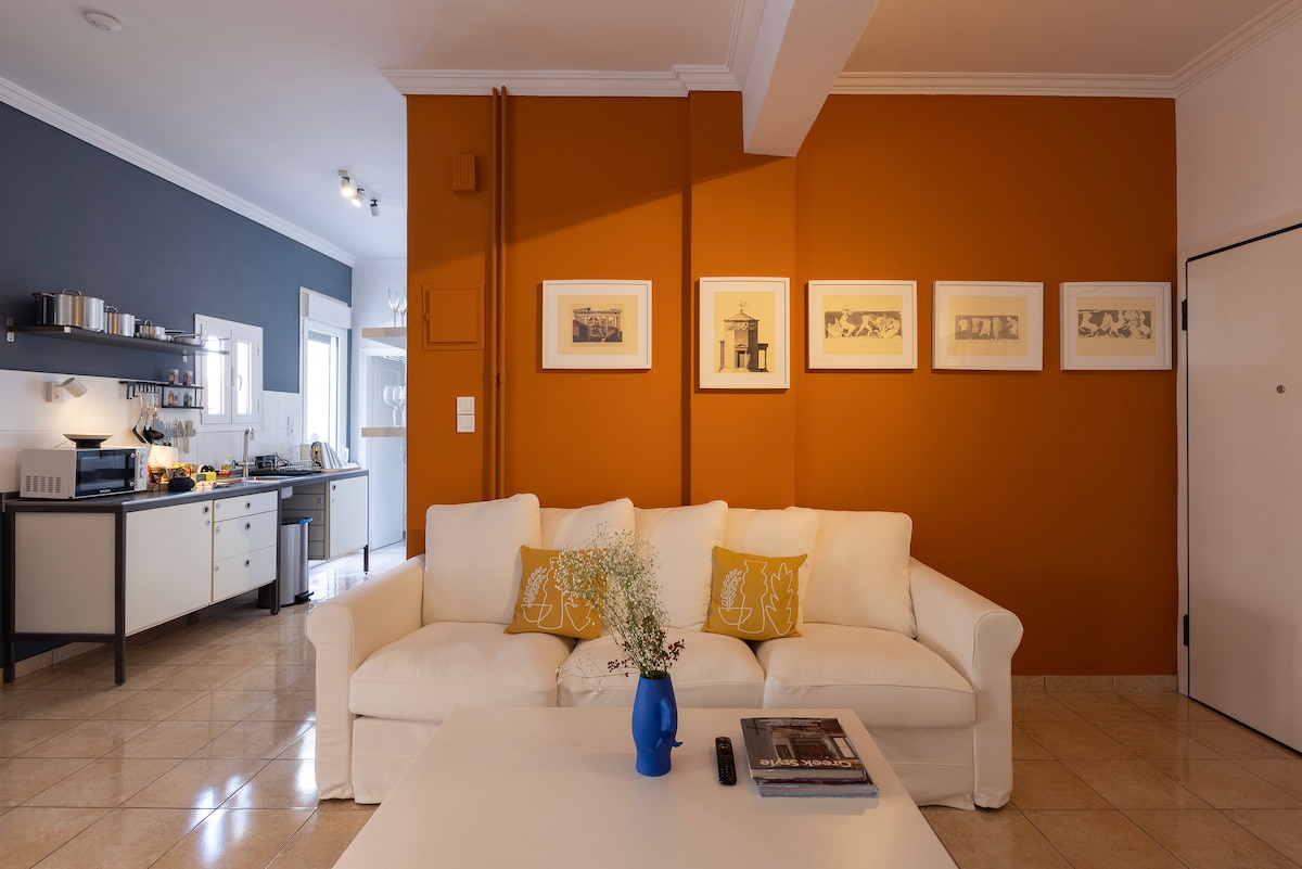 A living area showcases a light-colored sofa adorned with decorative pillows. A white coffee table is centered on a tiled floor. An accent wall in warm orange contrasts with framed artwork above the sofa. The kitchen can be seen in the background.