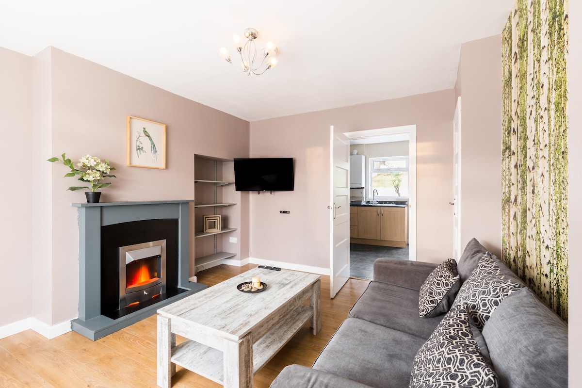 A living area features a cozy sofa adorned with patterned cushions, complemented by a rustic coffee table. A modern fireplace is positioned against a soft pink wall, while a large window provides natural light. A kitchen can be seen through an open doorway.