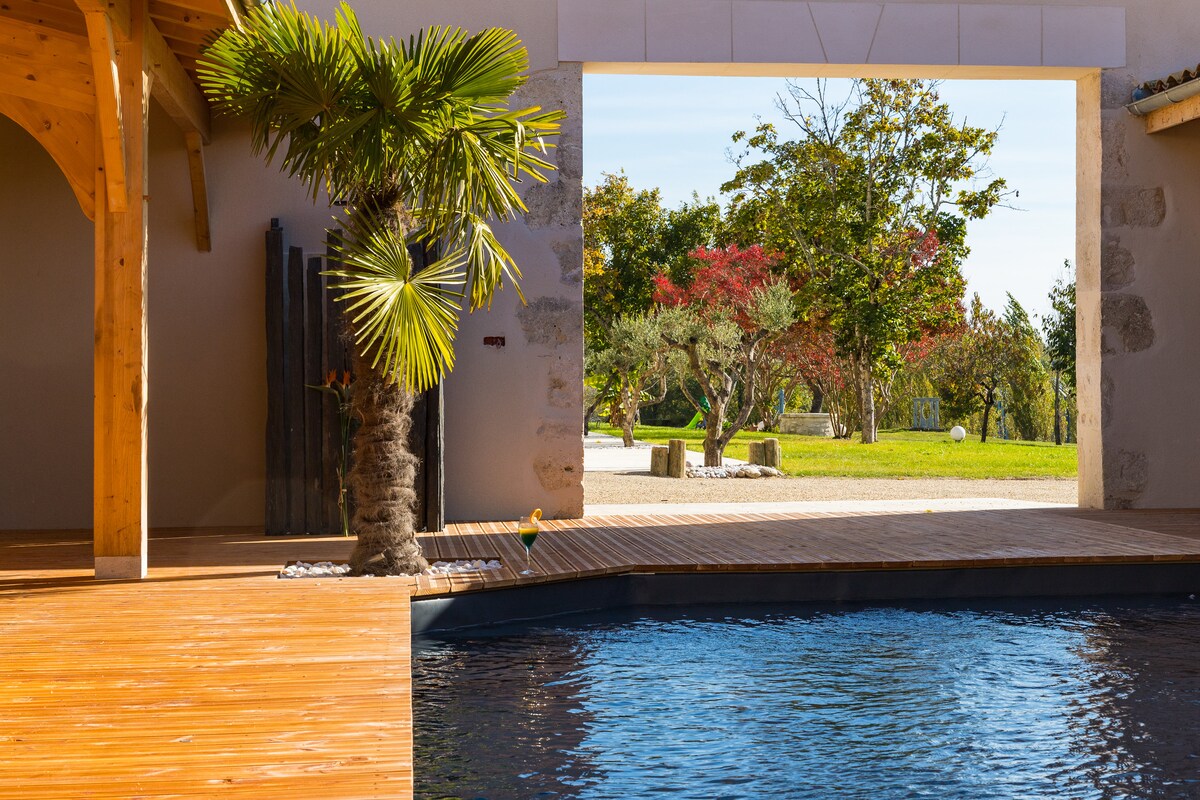 A serene view features an outdoor space framed by natural elements. A palm tree stands beside the pool, while large openings reveal a garden filled with trees and greenery, inviting a peaceful atmosphere.
