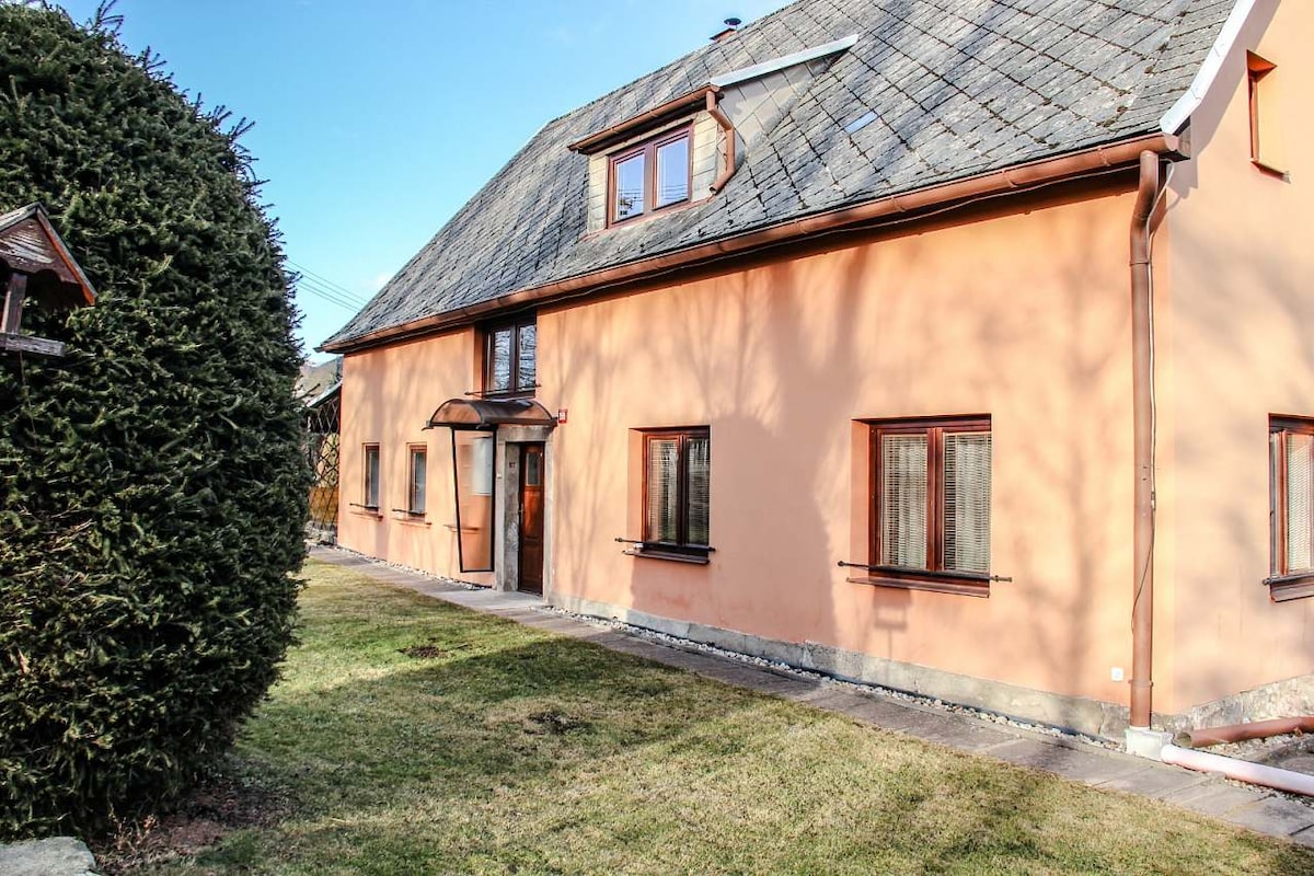 A two-story cottage with a soft peach exterior is framed by a neatly manicured lawn. The front entrance is highlighted by a simple archway and is flanked by several windows, providing a welcoming facade. A large evergreen shrub adds greenery to the setting.