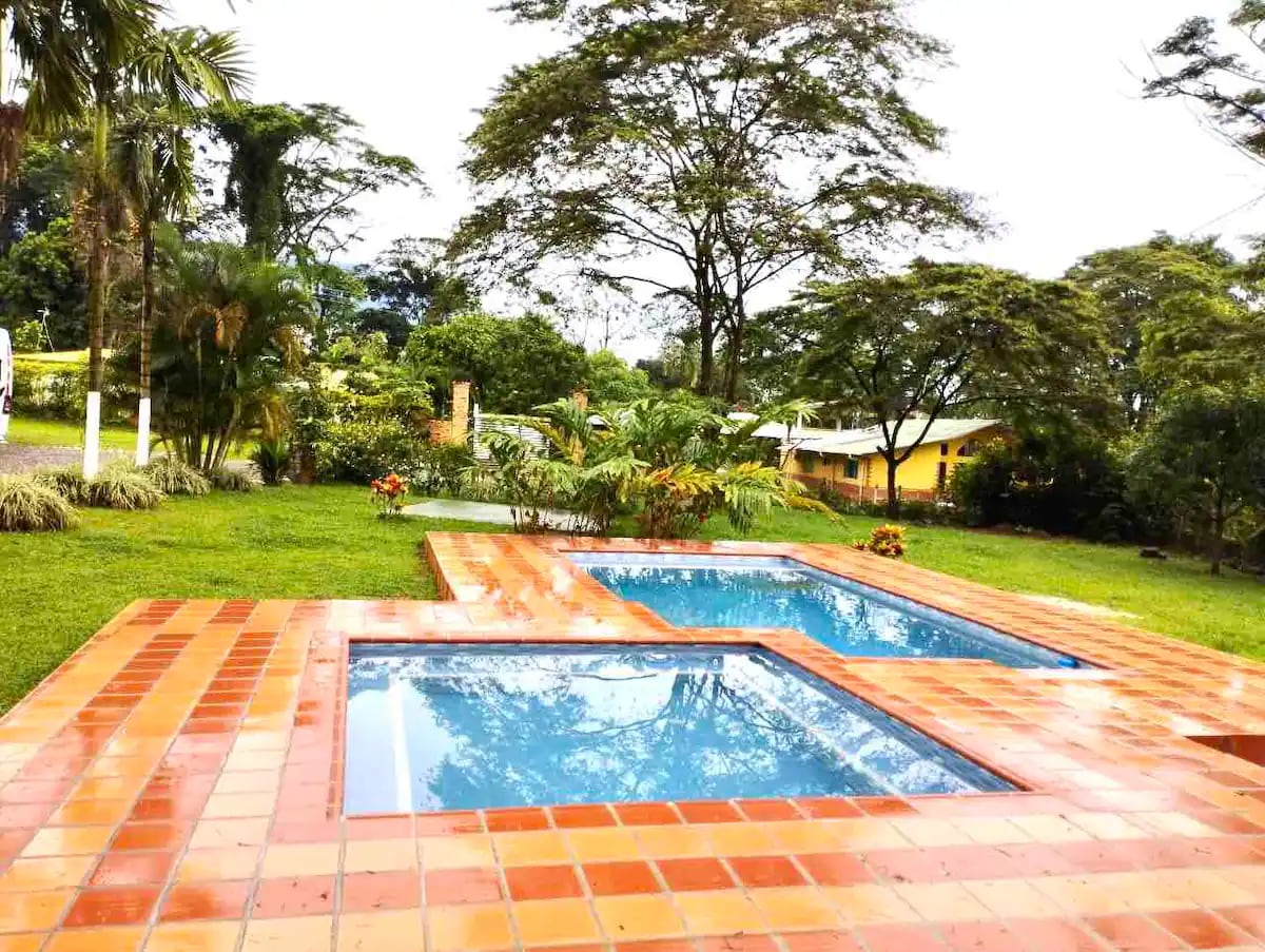 An outdoor area is depicted featuring two swimming pools set within terracotta tiles. Lush greenery surrounds the pools, with various plants and trees providing a natural backdrop. The vibrant colors of flowers add charm to the landscape, enhancing the serene atmosphere.