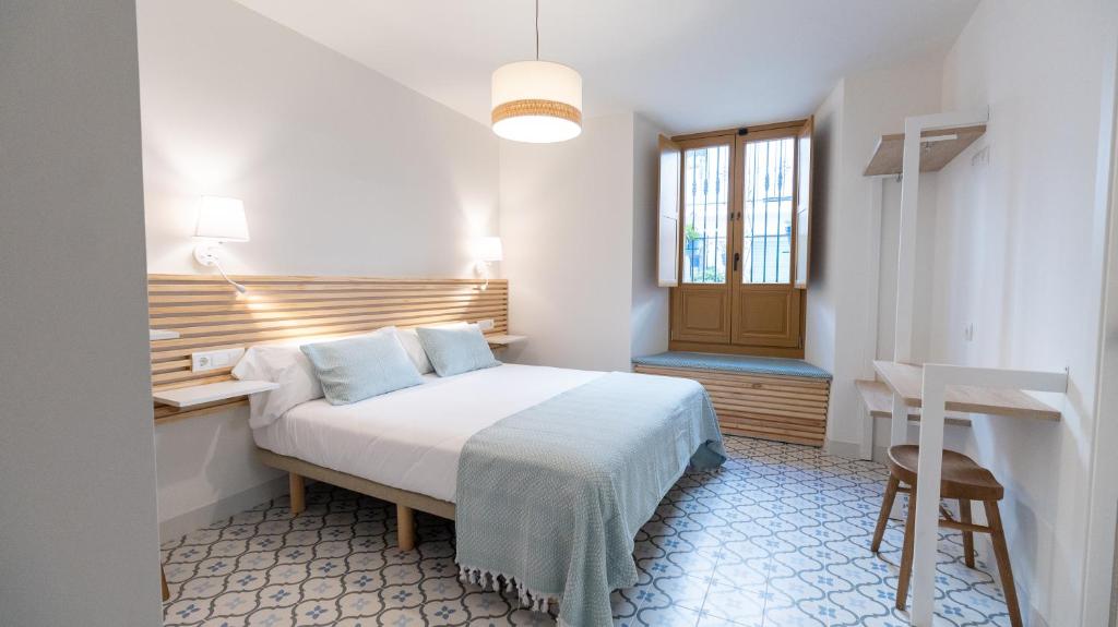 A spacious room is furnished with a large bed dressed in soft linens and complemented by light blue accents. Two wide windows allow natural light to fill the space, and unique tile flooring adds character. A wooden bench and a small stool provide additional seating.