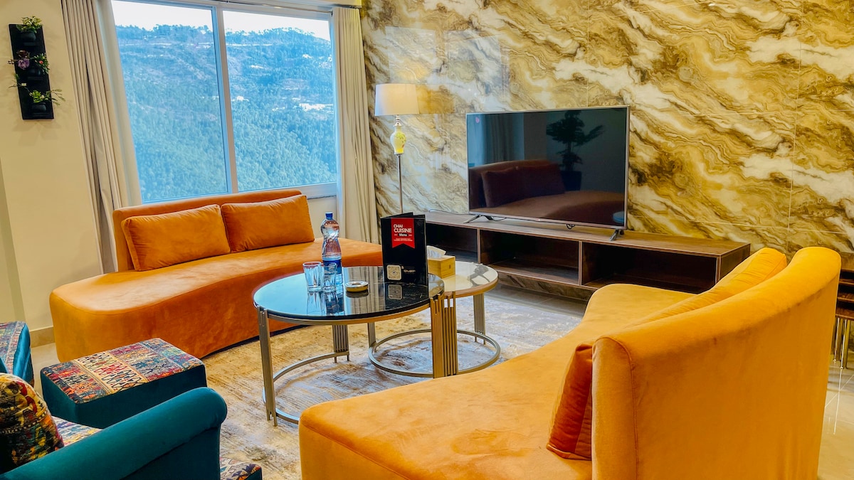 The living area features two orange curved sofas surrounding a glass coffee table. A flat-screen TV is mounted on a textured marble wall, offering a view of the mountains through a large window. Natural light brightens the space, creating a cheerful ambiance.
