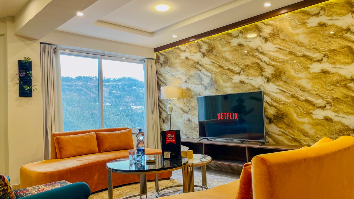A spacious living area features modern orange sofas arranged around a glass coffee table. A large television screen displaying Netflix is mounted against a textured, golden wall. Natural light streams through a window, offering a view of the surrounding mountains.