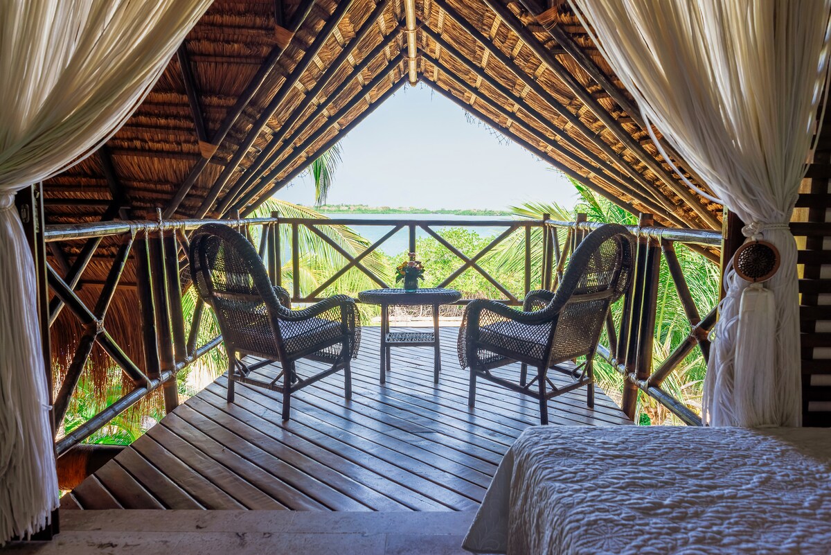 A spacious balcony area is featured, showcasing two wicker chairs facing a serene view of lush greenery and a body of water. The ceiling, adorned with natural materials, creates a cozy ambiance, while sheer drapes add an element of softness to the space.