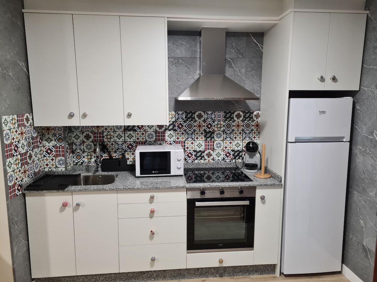A modern kitchen features sleek white cabinetry and a grey granite countertop. Colorful patterned tiles create visual interest on the backsplash. Appliances include a microwave, coffee maker, oven, and refrigerator, providing a fully equipped space for cooking and dining.