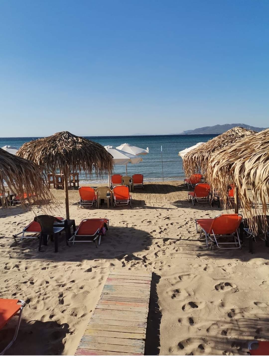 A sandy beach scene features rows of orange beach chairs beneath straw umbrellas. A wooden pathway leads through the sand toward the calm blue sea, with distant mountains visible on the horizon. The overall atmosphere suggests a relaxing coastal retreat.
