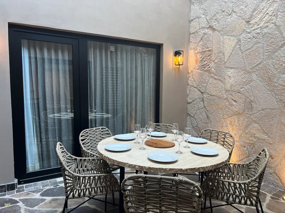 A round granite table is set for dining, surrounded by six woven chairs. Soft lighting highlights the textured stone wall, and large windows provide a view into the space. The atmosphere suggests a comfortable outdoor dining area.