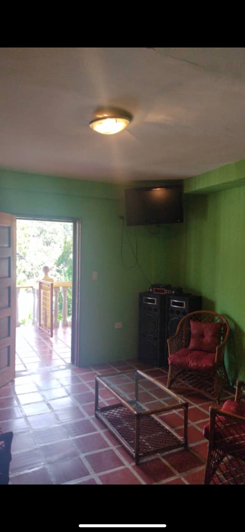 A living area features bright green walls and natural light from an open door leading to a balcony. A television is mounted on the wall above a pair of speakers, and a glass-top table is accompanied by two wicker chairs with red cushions.