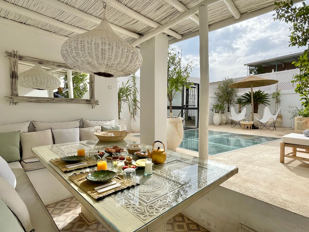 An inviting dining area showcases a glass-top table set with Moroccan dishes, surrounded by comfortable seating. Lush greenery and decorative elements are present, with a glimpse of a refreshing pool and outdoor lounge chairs in the background, all under a partially shaded area.