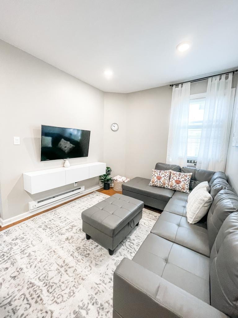 The living area features a modern gray sectional sofa arranged around a low ottoman and a flat-screen television mounted on a white console. Light-colored curtains frame a window, while a decorative clock and a small plant add subtle details to the space.