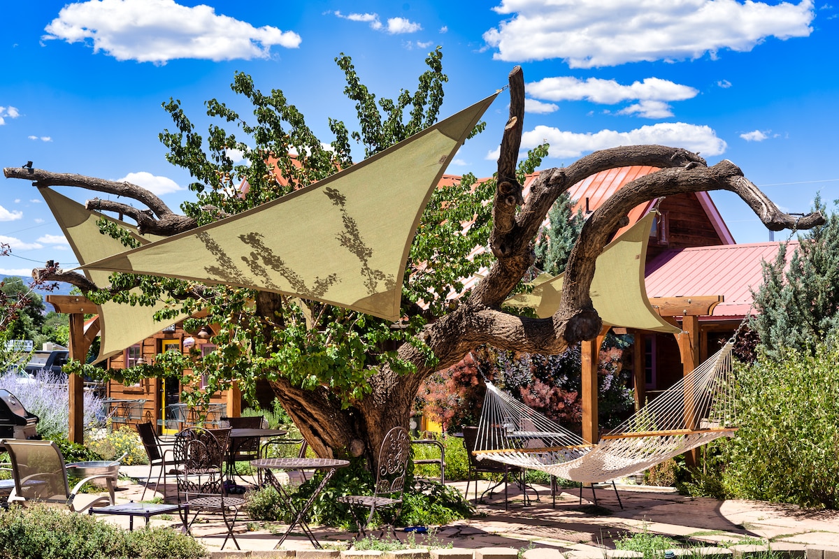 A large tree serves as a focal point with fabric canopies draping overhead, providing shade. Two hammocks are nestled between its branches, while outdoor seating arrangements are visible beneath, inviting relaxation in the garden area.