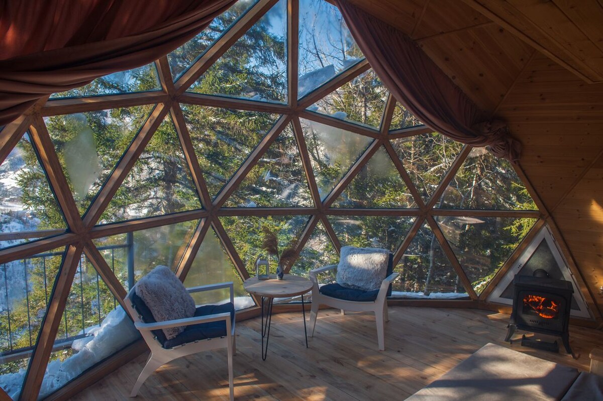 A unique geodesic dome interior features large triangular windows, offering views of surrounding trees. Two white armchairs with blue cushions are positioned near a small round table. A rustic stove is visible in one corner, enhancing the cozy atmosphere of the space.