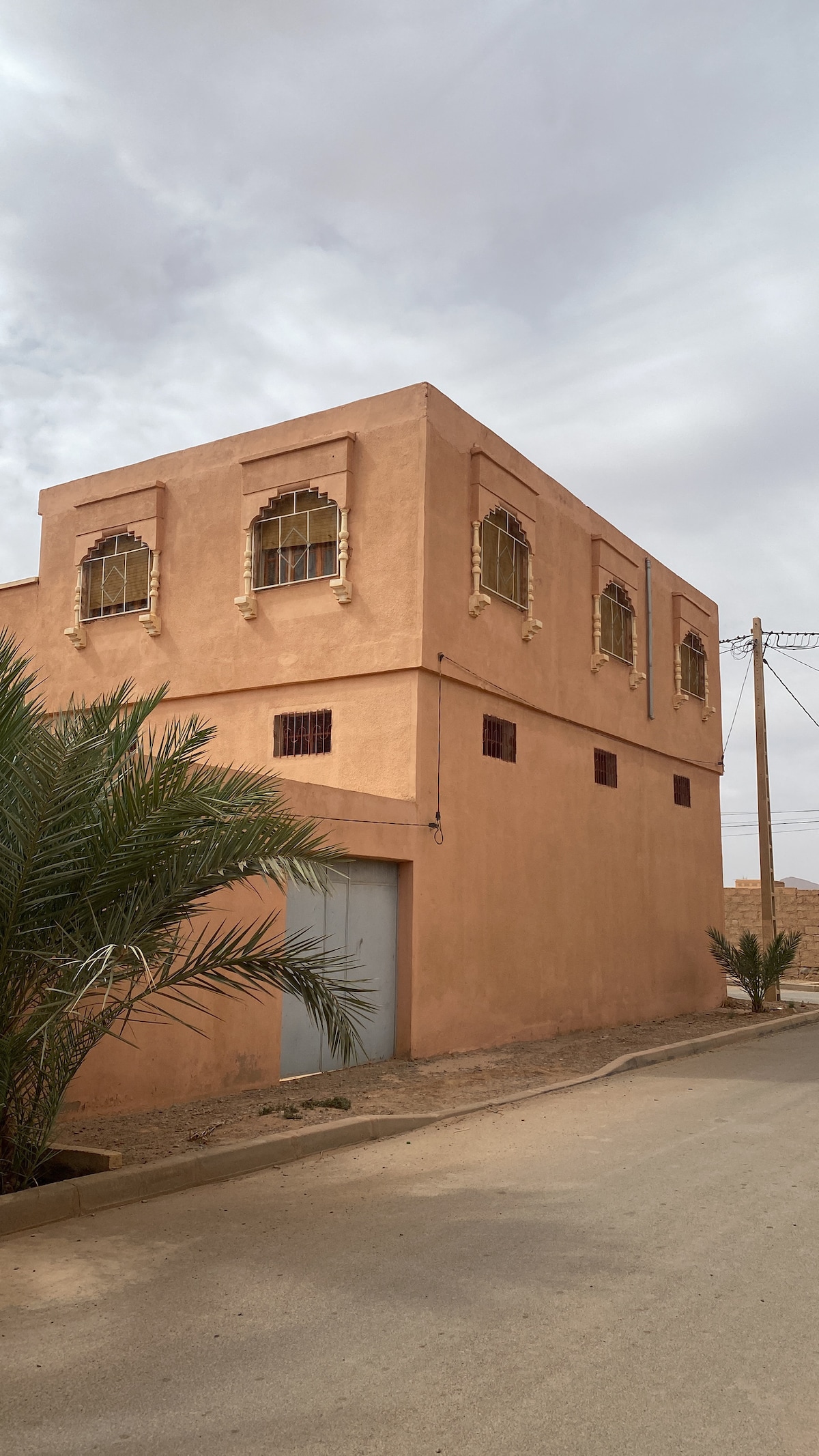 A two-story building is situated in a serene desert environment. The exterior features warm earthy tones, with arched windows framed in ornate detailing. Subtle palm trees are visible nearby, and a smooth road leads to the entrance, blending harmoniously with the surrounding landscape.