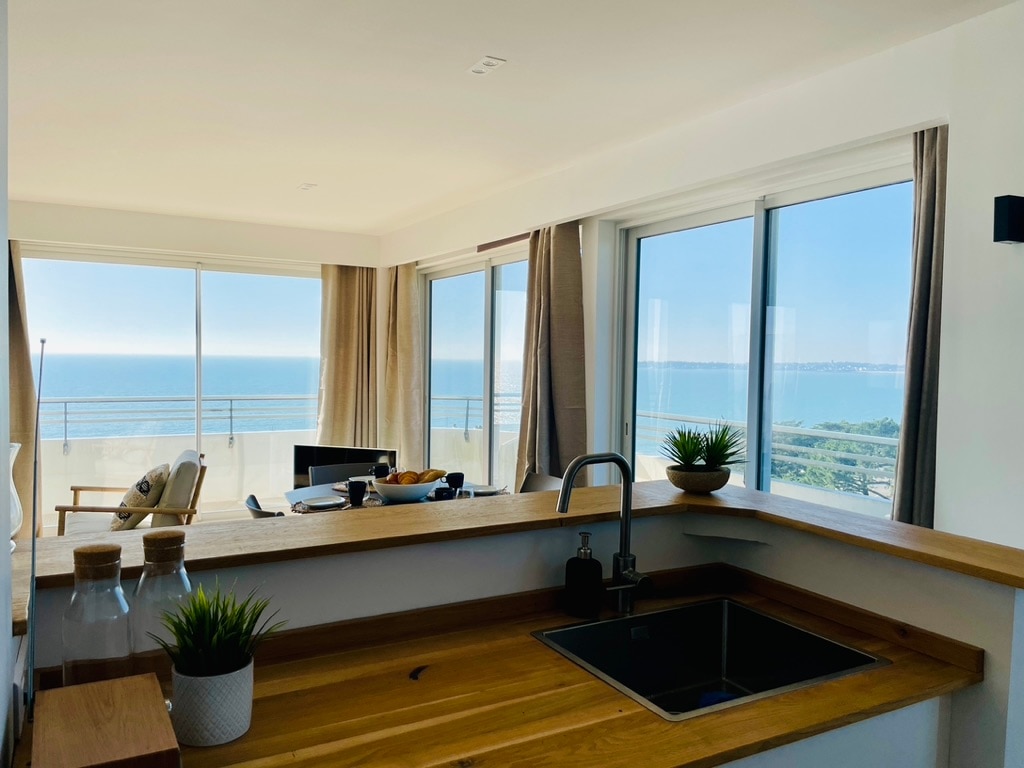 A modern open-plan living area features a well-equipped kitchen with a sleek sink and wooden counter. Large windows provide unobstructed views of the sea and illuminate the space, while light-colored curtains frame the glass. The serene atmosphere highlights a harmonious blend of indoor and outdoor living.