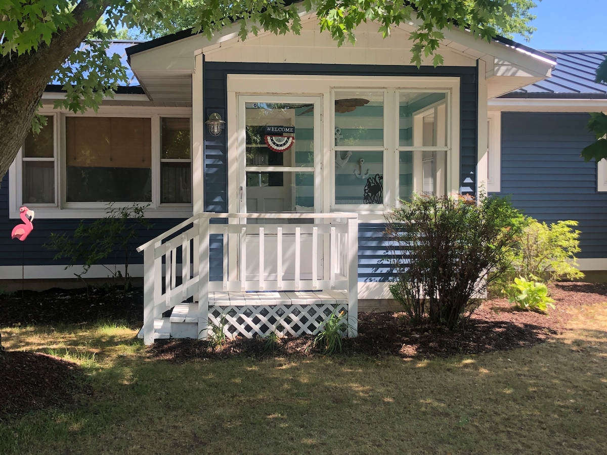A charming exterior view of the home showcases a blue facade accented with white trim. A small front porch features a white railing and steps leading to the lawn, bordered by leafy shrubs. A decorative welcome sign is visible through the window.