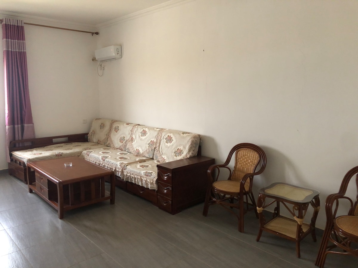 A spacious living area is furnished with a beige sofa adorned with floral patterns, accompanied by a wooden coffee table. Two rattan chairs are positioned beside a small side table, with soft natural light entering through the window, enhanced by light purple curtains.