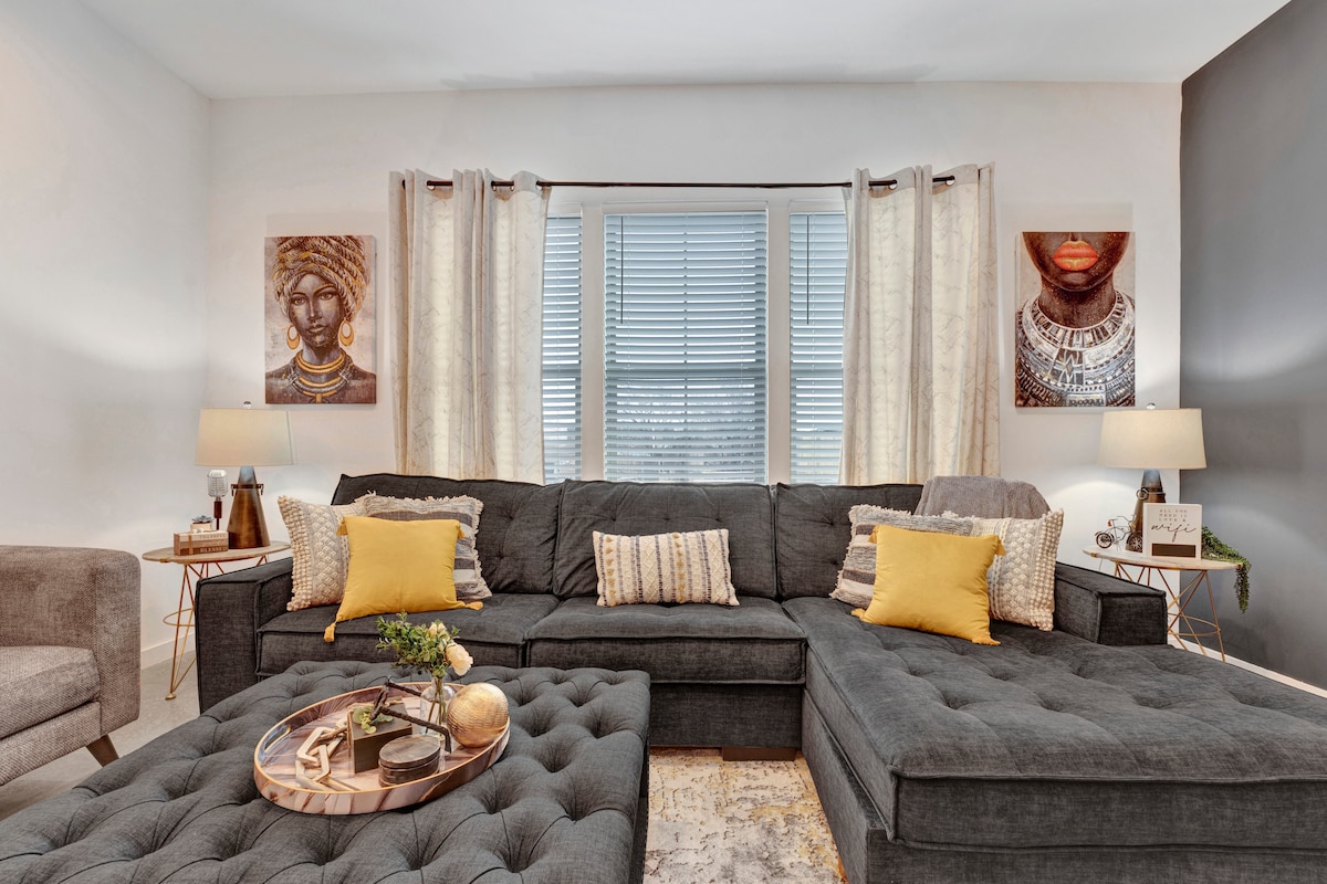 A modern living area features a large, dark sectional sofa adorned with yellow accent pillows. A coffee table with a decorative centerpiece sits in front of the sofa. Two art pieces hang on the walls, and light filters through the window, highlighting the soft drapery.