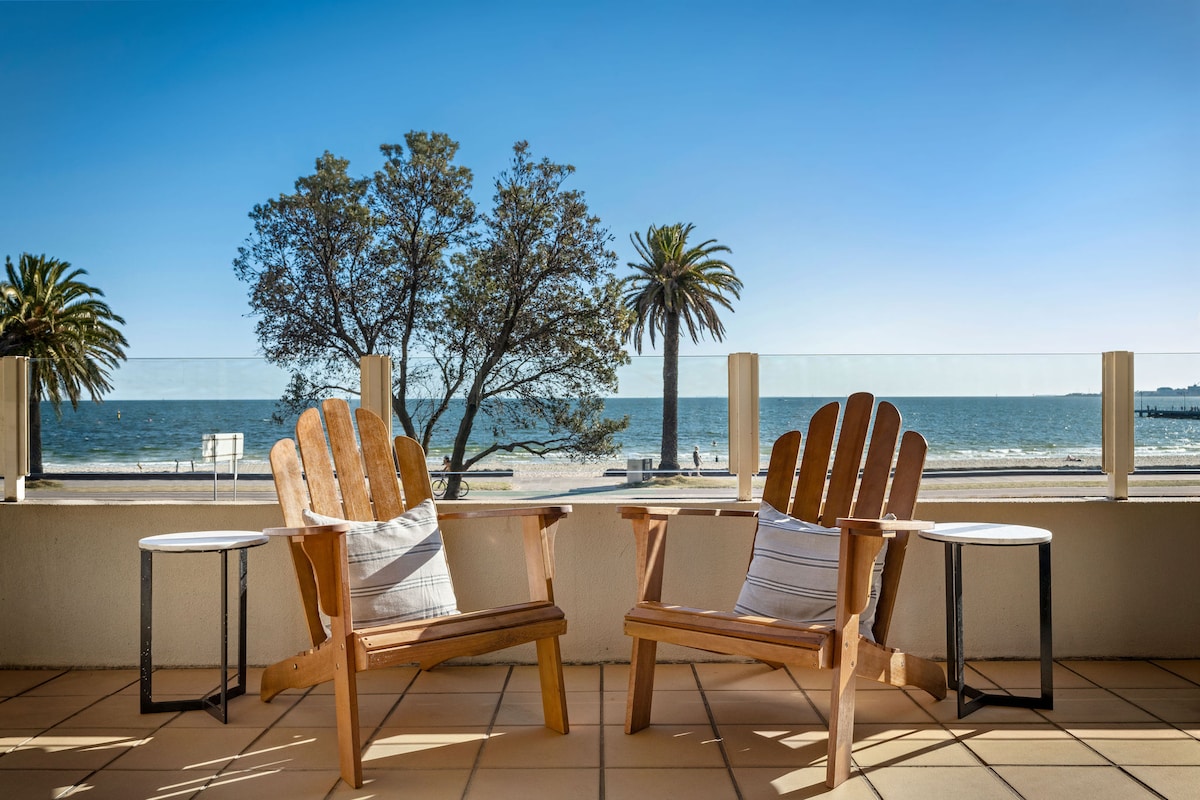 Two wooden chairs with cushions are positioned on a spacious terrace, overlooking the beach and bay. A clear glass railing offers unobstructed views of palm trees and the water. Small round tables are situated beside each chair, enhancing the inviting outdoor space.