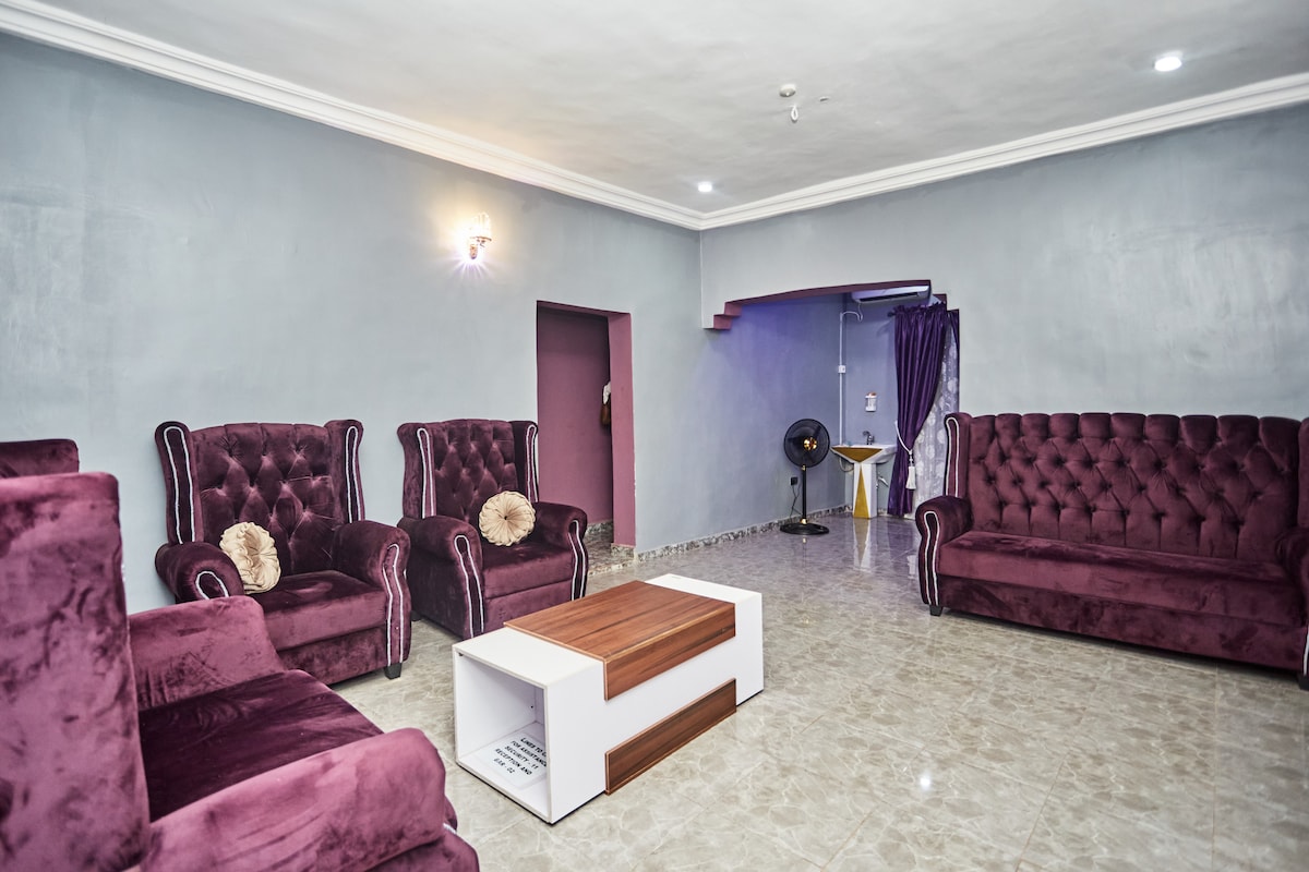 A spacious living area features plush, purple velvet sofas arranged around a wooden coffee table. The room is framed by light grey walls and includes a small, discreet fan in the corner. Soft lighting is provided by wall sconces, enhancing the comfort of the space.