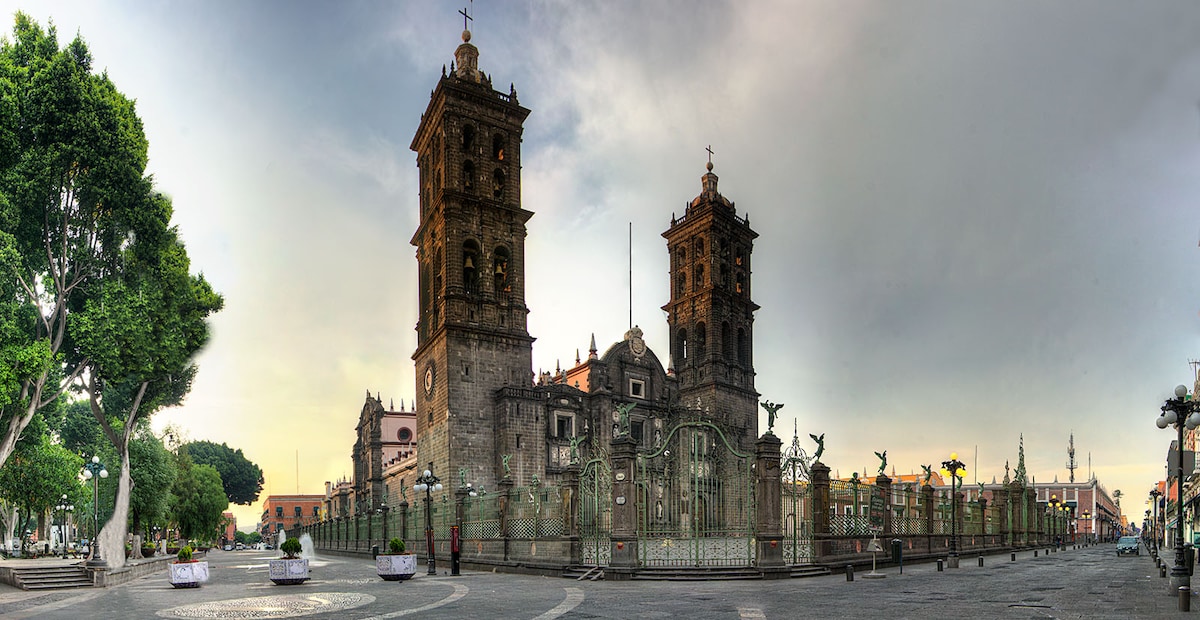 The historic Cathedral of Puebla is framed by lush greenery and a vibrant square. The two towering bell towers create a striking silhouette against a soft sky, while ornate gates and sculptures line the foreground, enhancing the architectural beauty of this cultural landmark.
