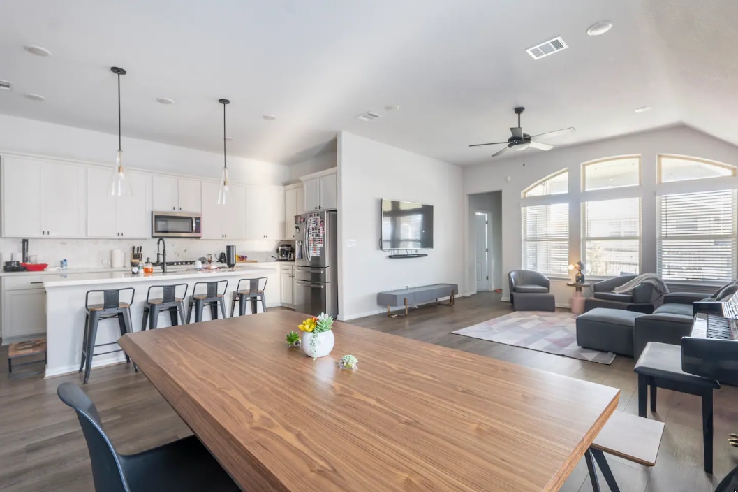 A bright and open living area showcases a large wooden dining table adorned with a small centerpiece. Modern kitchen fixtures and bar seating are visible in the background, with a cozy seating arrangement featuring dark sofas and a wall-mounted TV. Natural light floods the space through arched windows.