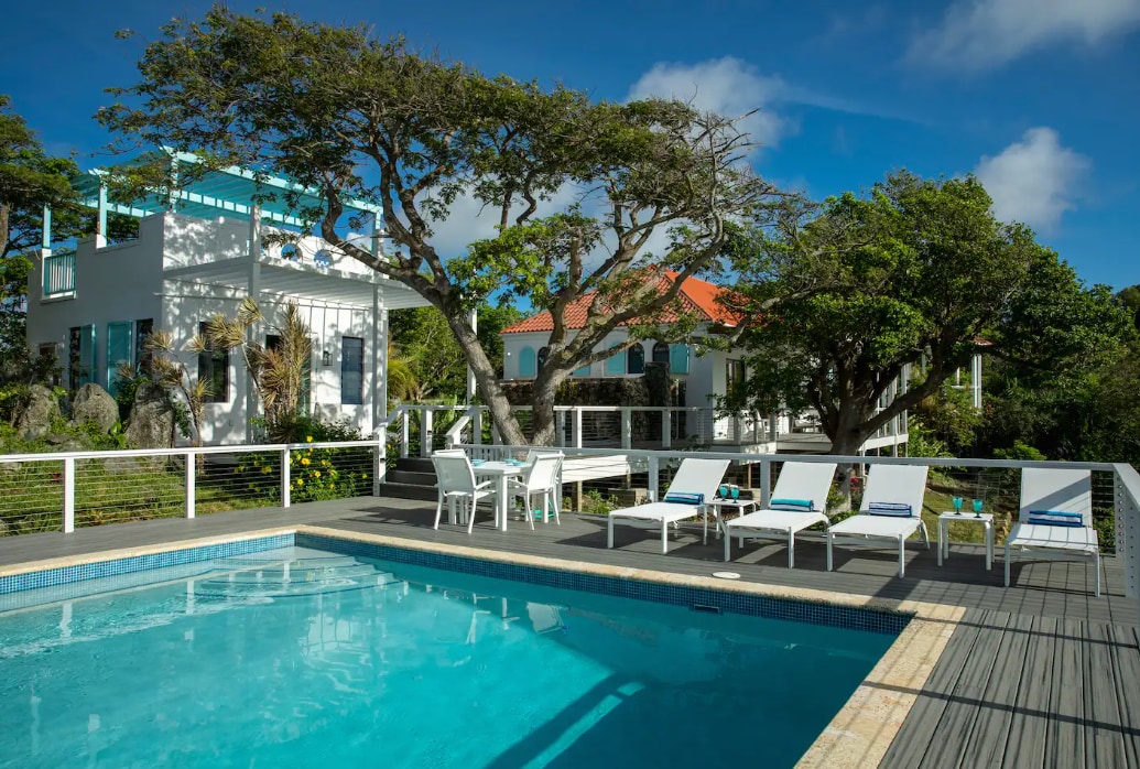 A serene pool area is surrounded by lounge chairs and lush greenery, offering a refreshing space for relaxation. The modern villa structure features a light color scheme and large windows, allowing for ample natural light and views of the landscape.