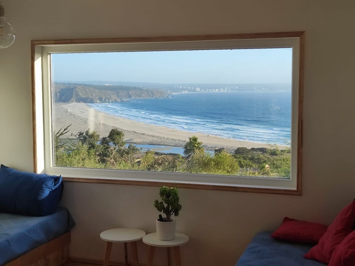 A large window frames a stunning view of the ocean and coastline. Waves can be seen hitting the sandy beach, complemented by a clear blue sky. Two small tables with a potted plant are positioned in front of the window, inviting relaxation.