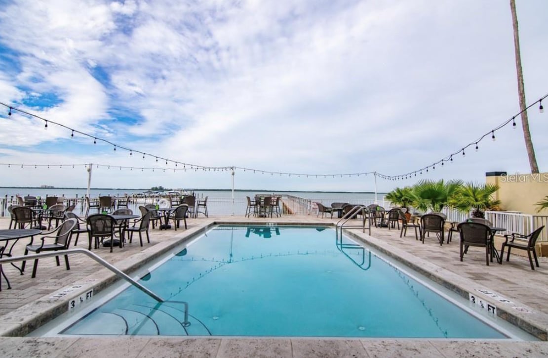 A refreshing pool area is framed by multiple seating options and string lights overhead. The clear water reflects the sky, with beach views visible in the background. Decorative landscaping elements enhance the ambiance, creating a relaxing environment for guests.