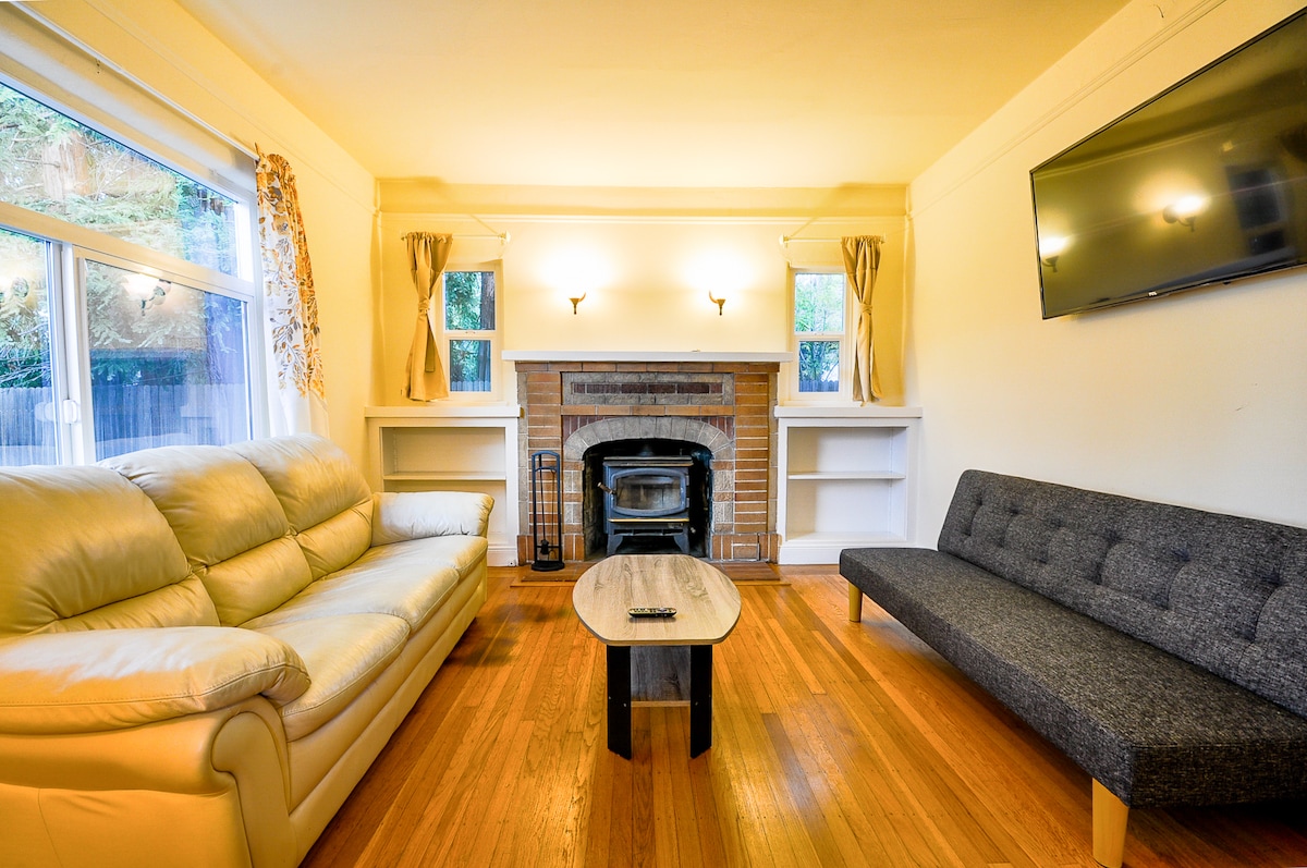 A living area showcases a light-colored leather sofa and a dark gray futon, arranged around a central coffee table. Large windows allow natural light to fill the space, while a brick fireplace adds warmth. A flat-screen TV is mounted on the wall.
