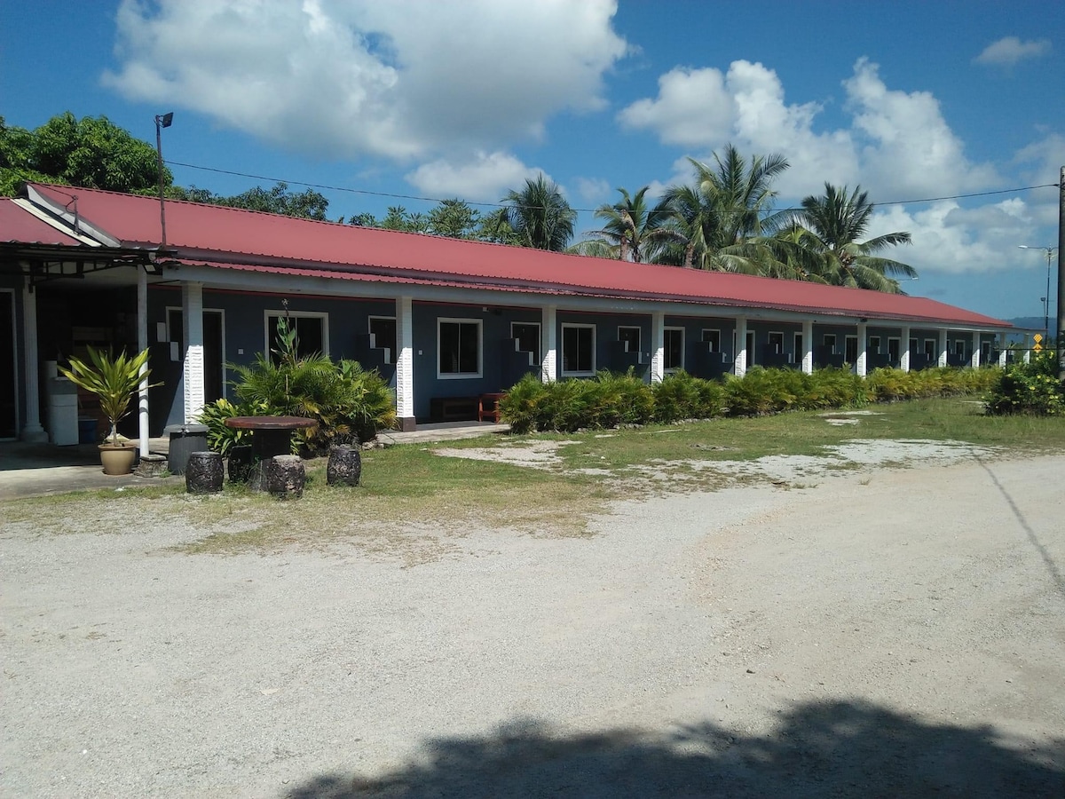 A row of single-story accommodations is visible beneath a clear blue sky. Lush greenery outlines the pathway, with tropical plants thoughtfully placed in pots. Each unit features a covered entry and large windows, creating an inviting façade that blends with the surrounding nature.