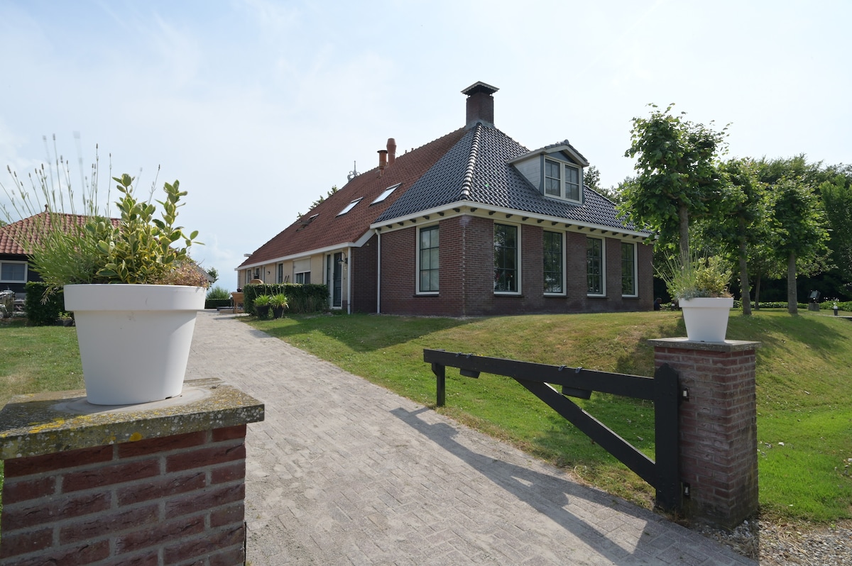 A red-brick house stands prominently against a clear sky, framed by well-maintained grassy areas. A decorative white planter sits on the stone wall, with an inviting pathway leading towards the entrance. The structure features multiple gabled roofs and large windows enhancing natural light.
