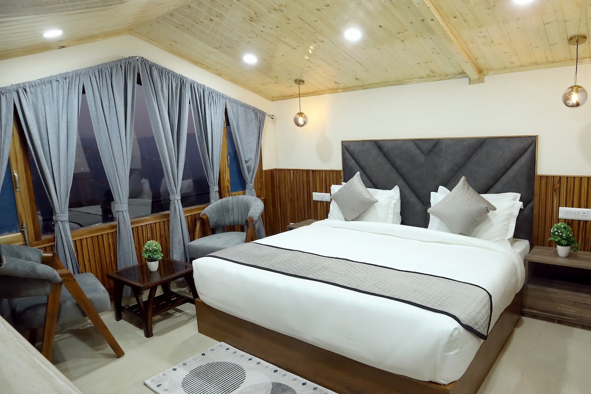 A well-appointed bedroom features a spacious bed with neatly arranged pillows. Large windows are draped with soft grey curtains, allowing natural light to brighten the room. Two stylish chairs complement a wooden nightstand, enhancing the room's modern design.