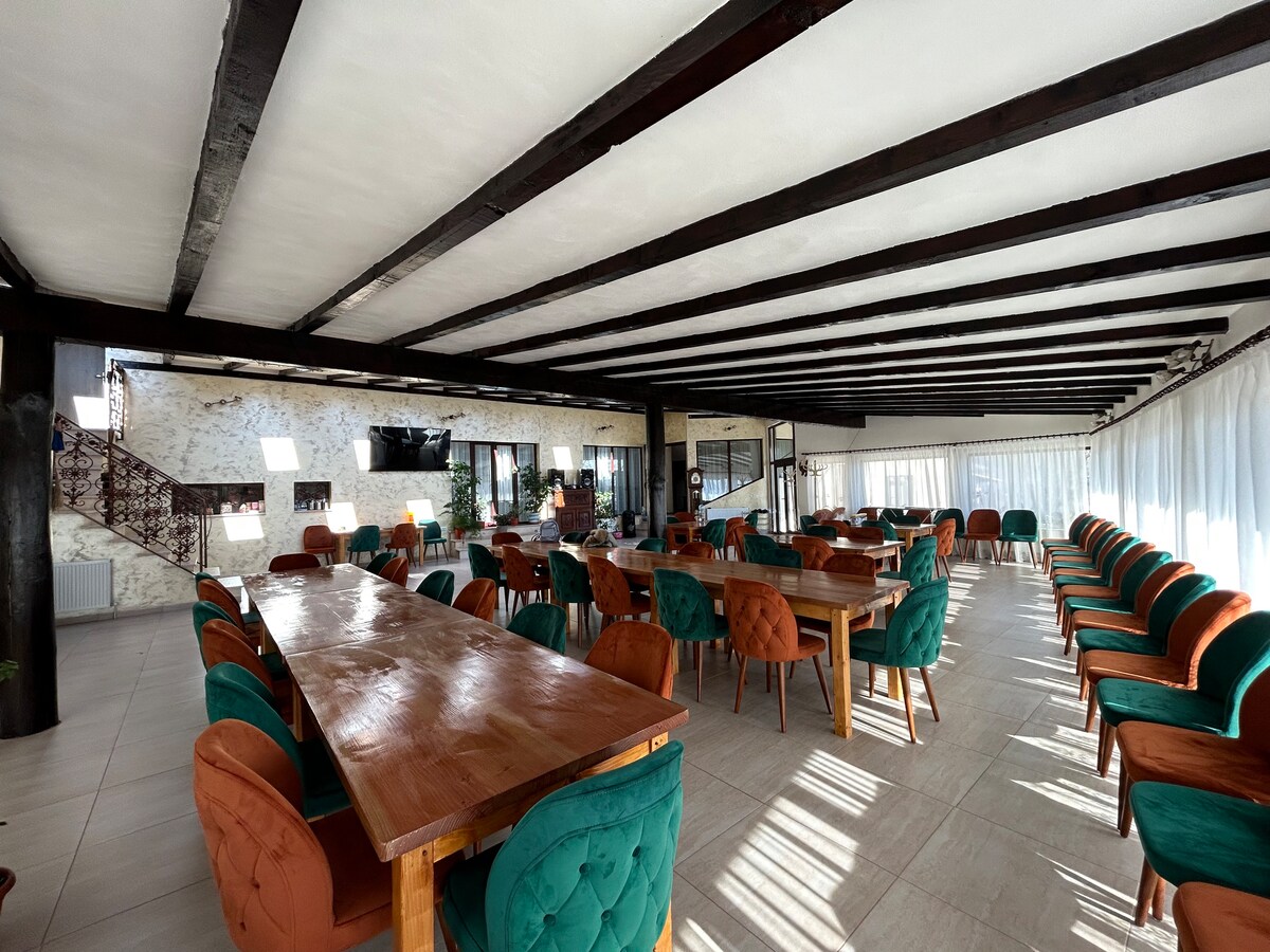 A spacious dining area is furnished with wooden tables and a row of comfortable chairs featuring green and orange upholstery. Exposed wooden beams form a striking visual overhead, while natural light filters through large windows adorned with sheer curtains.