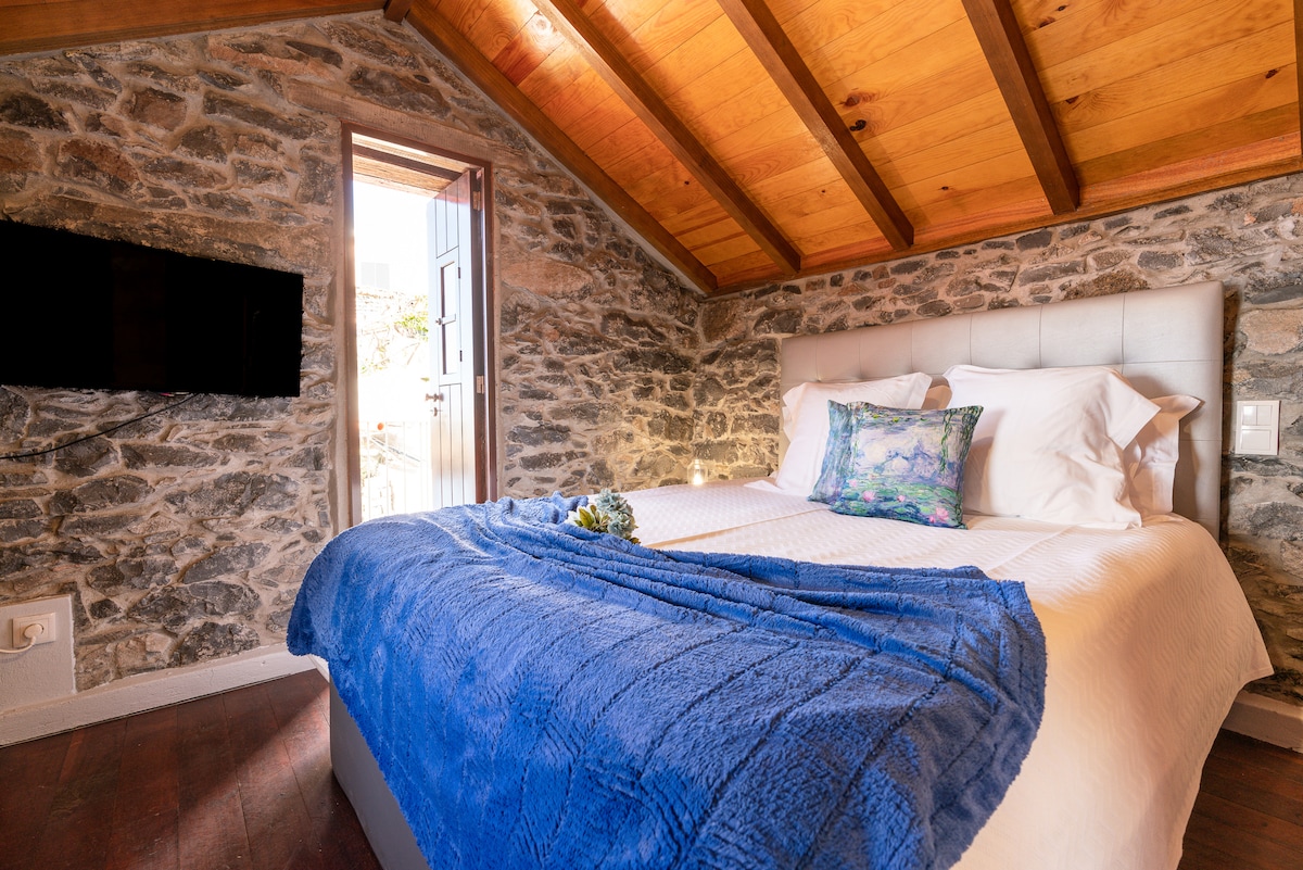 A cozy sleeping area is highlighted by a spacious bed adorned with a soft blue throw. Exposed stone walls add texture and warmth, while wooden beams stretch across the ceiling. Natural light enters through an open door, creating an inviting ambiance.