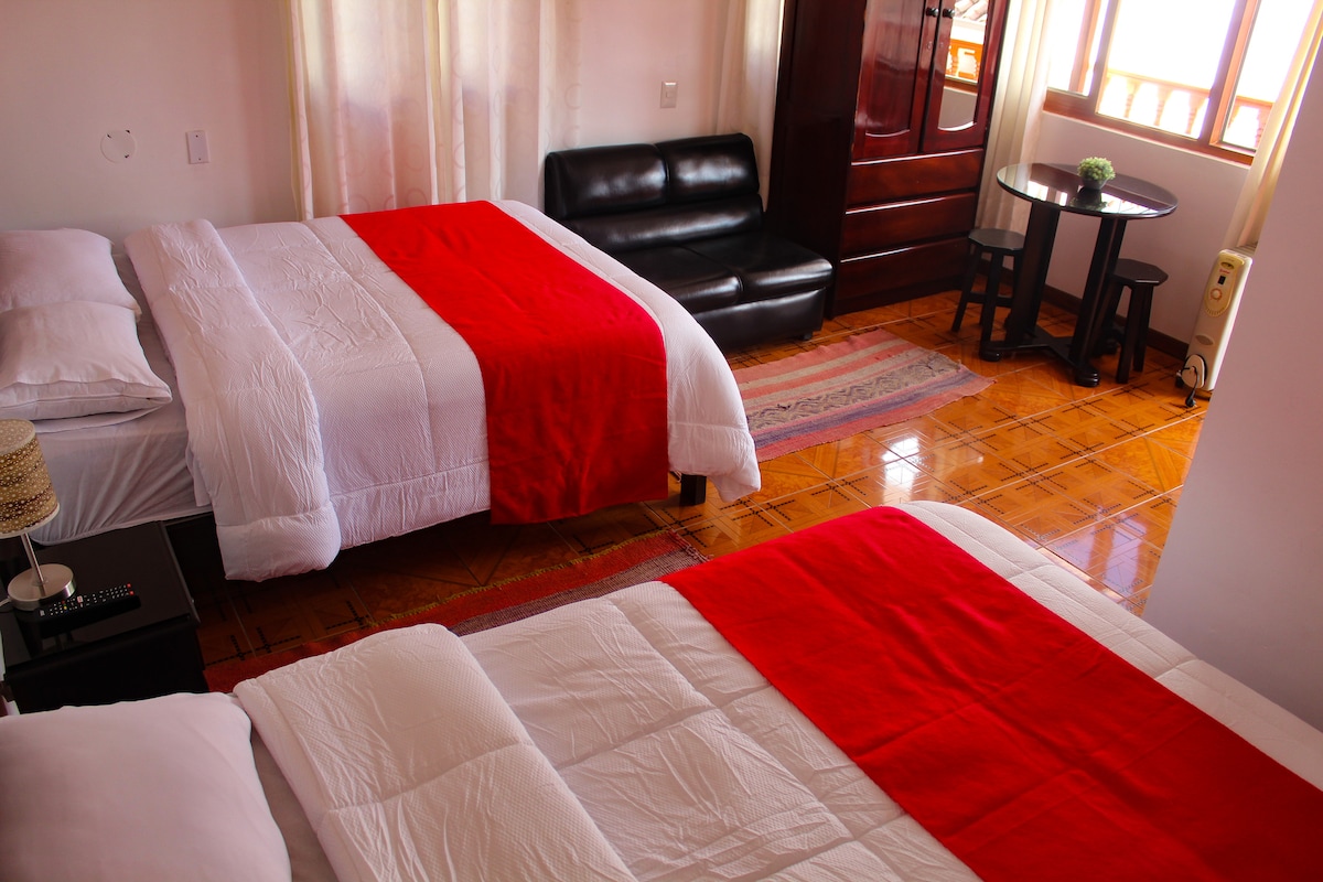 A spacious room is furnished with two beds, each topped with a red bedspread. A black sofa is positioned near a small table, creating a comfortable seating area. Large windows allow natural light to brighten the space, while hardwood flooring adds warmth.