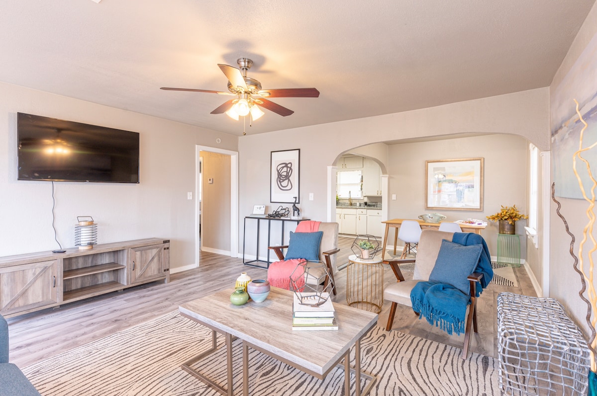 A spacious living area features soft-toned walls and a ceiling fan. Comfortable seating options are arranged around a central coffee table. A large flat-screen TV is mounted on the wall. Light streams through an archway leading to the kitchen, enhancing the open layout.