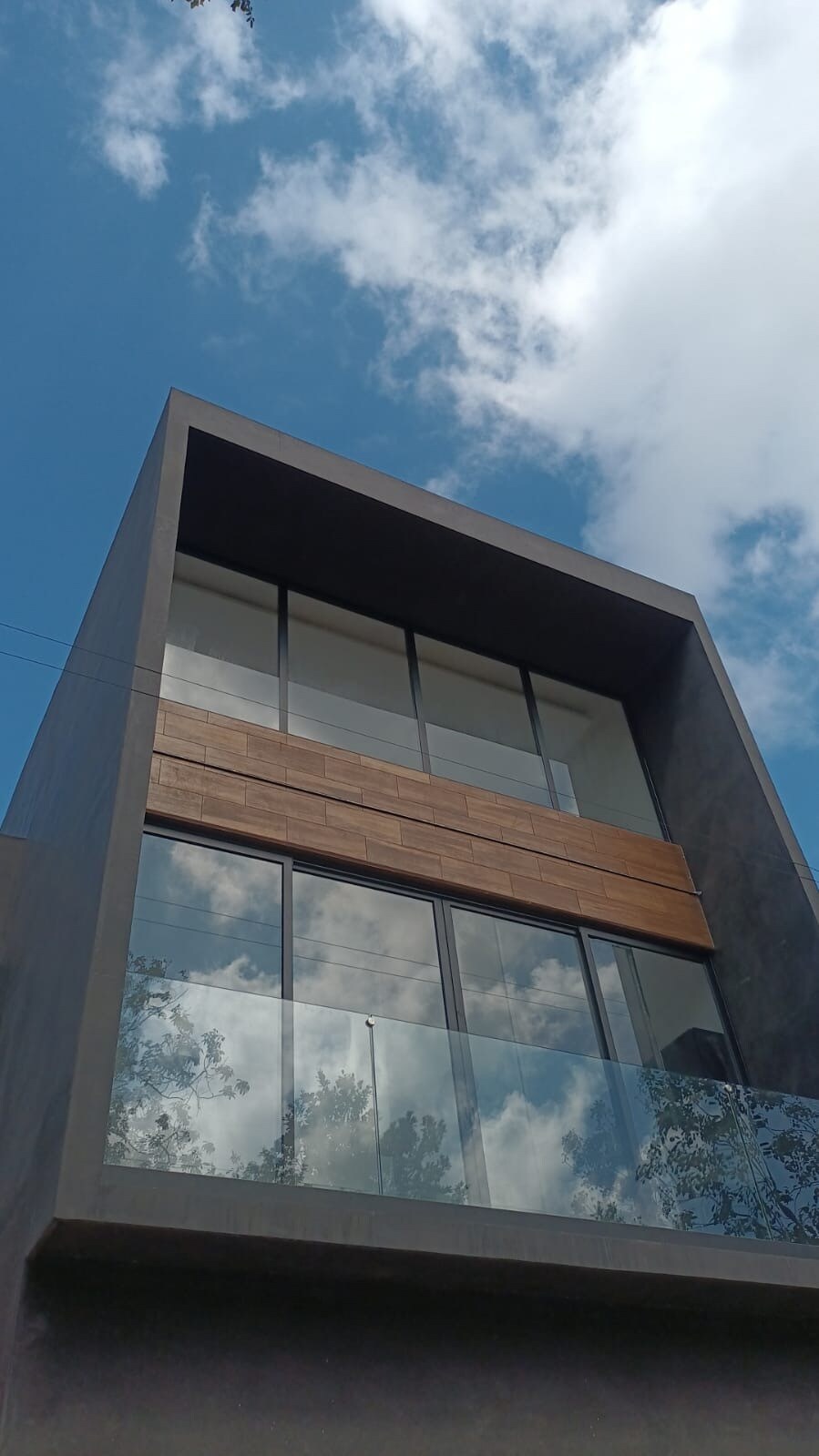 A modern structure features a sleek exterior with large glass windows that reflect the sky above. A wooden accent area is situated on the second level, providing a natural contrast to the contemporary design. The balcony offers a spacious, open feel.
