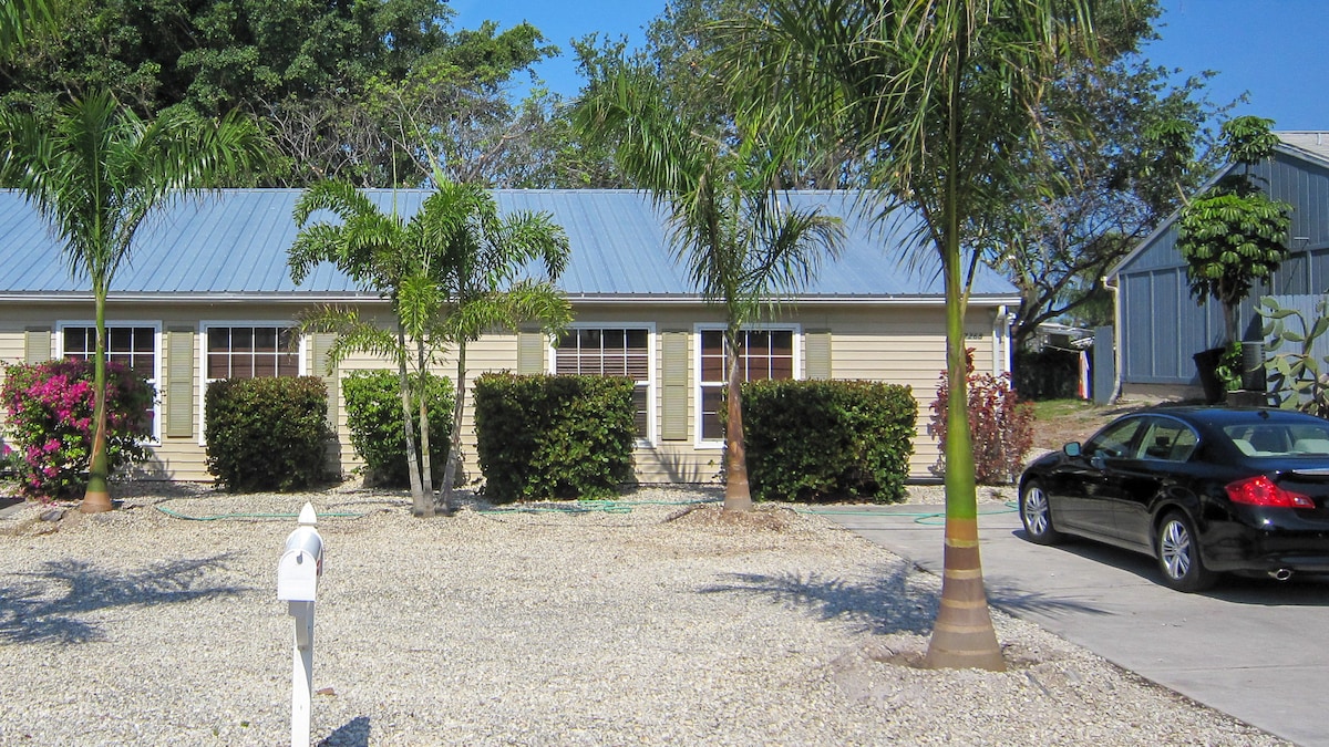 A light-colored single-story home features multiple windows framed by lush greenery and flowering plants. A gravel driveway is seen leading to a black car parked adjacent to the house. Palm trees provide shade in the front garden area.