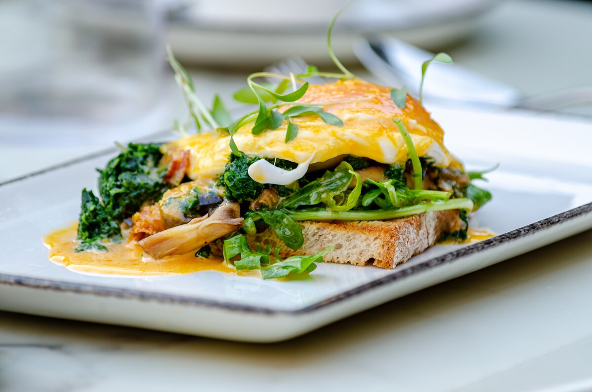 A plated dish showcases a slice of toasted bread topped with sautéed greens and a poached egg, drizzled with a creamy sauce. Fresh herbs garnish the dish, adding a vibrant touch to the presentation.